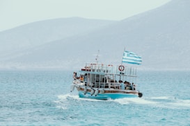A boat filled with passengers travels across a calm sea with mountains in the background. A Greek flag waves at the stern of the boat, and several people are gathered on deck, enjoying the ride.