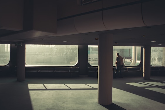 A spacious, dimly lit room with large floor-to-ceiling windows casting elongated shadows across the floor. A lone person is walking near one of the windows, creating a contemplative and solitary atmosphere. The design is modern, with smooth surfaces and a muted color palette.