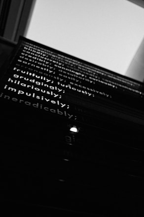 A monochrome image showcasing a series of words on a wall installation, viewed from a low angle, creating a sense of depth and perspective. The composition is focused on the words 'fruitfully,' 'furiously,' 'hilariously,' 'impulsively,' and 'ineradicably,' among others, layered in a poetic sequence.