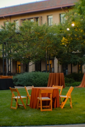 A grassy outdoor setting with wooden chairs and round tables covered in orange tablecloths. The backdrop features a building with a red-tiled roof and windows, partially obscured by lush green trees. The area appears calm and prepared for an event.