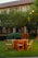A grassy outdoor setting with wooden chairs and round tables covered in orange tablecloths. The backdrop features a building with a red-tiled roof and windows, partially obscured by lush green trees. The area appears calm and prepared for an event.