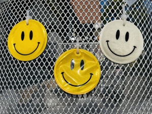 Three smiley face air fresheners hang on a metallic mesh surface, with two in yellow and one in white. The background contains blurred reflections and a hint of greenery.