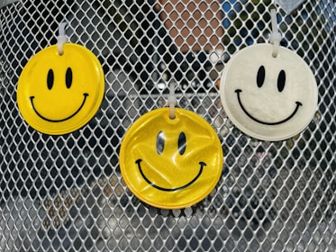 Three smiley face air fresheners hang on a metallic mesh surface, with two in yellow and one in white. The background contains blurred reflections and a hint of greenery.