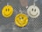 Three smiley face air fresheners hang on a metallic mesh surface, with two in yellow and one in white. The background contains blurred reflections and a hint of greenery.