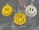 Three smiley face air fresheners hang on a metallic mesh surface, with two in yellow and one in white. The background contains blurred reflections and a hint of greenery.