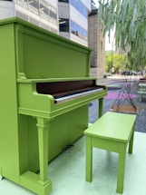 A bright green upright piano accompanied by a matching bench placed outdoors on a city street. Surrounding urban elements include buildings in the background, a tree sculpture, and colorful patterns on the ground.