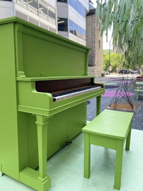 A bright green upright piano accompanied by a matching bench placed outdoors on a city street. Surrounding urban elements include buildings in the background, a tree sculpture, and colorful patterns on the ground.