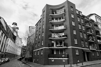 A black and white image captures an urban scene featuring a tall, curved apartment building. The structure is constructed with brick and has several balconies. On the left side, a narrower, industrial-style building with large fans or chimneys is visible. A paved street runs between the buildings, lined with parked cars, and a bare tree stands near the apartment.