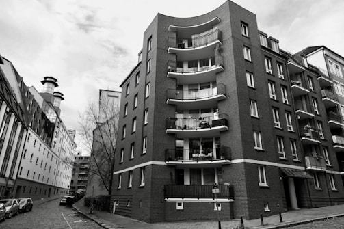 A black and white image captures an urban scene featuring a tall, curved apartment building. The structure is constructed with brick and has several balconies. On the left side, a narrower, industrial-style building with large fans or chimneys is visible. A paved street runs between the buildings, lined with parked cars, and a bare tree stands near the apartment.