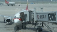 A commercial airplane is parked at an airport gate connected to a boarding bridge with visible branding on the side. Ground crew and vehicles are visible on the tarmac, with another plane in the background.