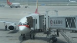 A commercial airplane is parked at an airport gate connected to a boarding bridge with visible branding on the side. Ground crew and vehicles are visible on the tarmac, with another plane in the background.