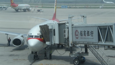 A commercial airplane is parked at an airport gate connected to a boarding bridge with visible branding on the side. Ground crew and vehicles are visible on the tarmac, with another plane in the background.