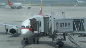 A commercial airplane is parked at an airport gate connected to a boarding bridge with visible branding on the side. Ground crew and vehicles are visible on the tarmac, with another plane in the background.