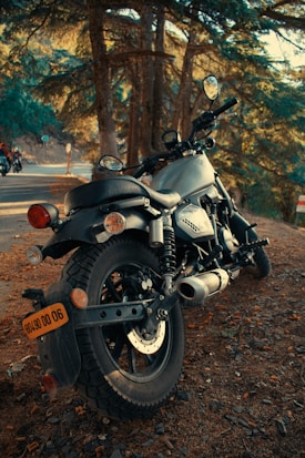 A motorcycle is parked on a forested roadside with sunlight filtering through the trees. The focus is on the rear wheel and exhaust pipes, with the setting creating a serene and adventurous atmosphere. The road curves slightly, and another motorcycle is visible in the distance.