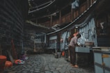 A person is washing dishes at an outdoor sink in a traditional, round, multi-story building with wooden and brick structures. Various household items like brooms, buckets, and stools are scattered around the stone-paved courtyard. Laundry hangs from the upper level railings of the building.