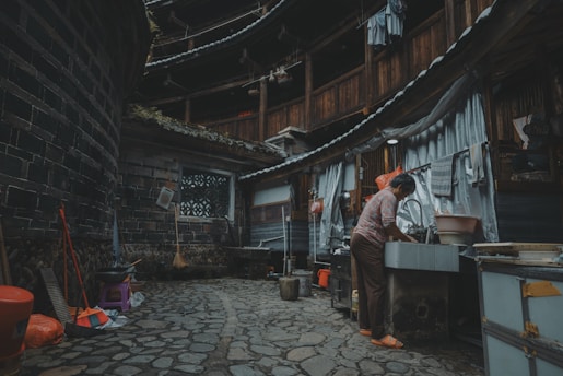 A person is washing dishes at an outdoor sink in a traditional, round, multi-story building with wooden and brick structures. Various household items like brooms, buckets, and stools are scattered around the stone-paved courtyard. Laundry hangs from the upper level railings of the building.