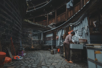 A person is washing dishes at an outdoor sink in a traditional, round, multi-story building with wooden and brick structures. Various household items like brooms, buckets, and stools are scattered around the stone-paved courtyard. Laundry hangs from the upper level railings of the building.