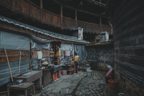 A rustic outdoor kitchen setup in a courtyard area with stone flooring. The walls are made of wooden panels and bricks, partially covered with corrugated material. Various kitchen utensils, pots, and containers are scattered around, with a person wearing a yellow jacket engaged in cooking or cleaning.