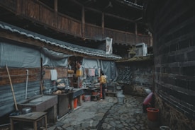 A rustic outdoor kitchen setup in a courtyard area with stone flooring. The walls are made of wooden panels and bricks, partially covered with corrugated material. Various kitchen utensils, pots, and containers are scattered around, with a person wearing a yellow jacket engaged in cooking or cleaning.