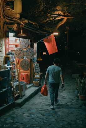 A person walks down a cobblestone path at night, carrying a red shopping bag. They are passing a small shop adorned with traditional Chinese decorations and a Chinese flag. The shop is lit up, with various goods displayed outside, including ornate, circular wooden carvings and other souvenirs. The surrounding area is dimly lit with foliage extending over the path.