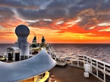 A cruise ship deck overlooking a vast ocean during sunset. The sky is ablaze with vibrant shades of orange, red, and yellow, blending into the darker hues of approaching night. A large radar sphere is prominently featured in the foreground, and the ship's layout includes seating and a water slide. Smoke trails rise from dual exhaust stacks, adding to the serene yet dynamic scene.