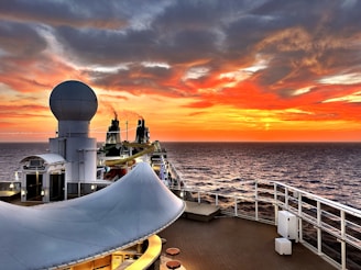 A cruise ship deck overlooking a vast ocean during sunset. The sky is ablaze with vibrant shades of orange, red, and yellow, blending into the darker hues of approaching night. A large radar sphere is prominently featured in the foreground, and the ship's layout includes seating and a water slide. Smoke trails rise from dual exhaust stacks, adding to the serene yet dynamic scene.