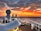 A cruise ship deck overlooking a vast ocean during sunset. The sky is ablaze with vibrant shades of orange, red, and yellow, blending into the darker hues of approaching night. A large radar sphere is prominently featured in the foreground, and the ship's layout includes seating and a water slide. Smoke trails rise from dual exhaust stacks, adding to the serene yet dynamic scene.