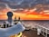 A cruise ship deck overlooking a vast ocean during sunset. The sky is ablaze with vibrant shades of orange, red, and yellow, blending into the darker hues of approaching night. A large radar sphere is prominently featured in the foreground, and the ship's layout includes seating and a water slide. Smoke trails rise from dual exhaust stacks, adding to the serene yet dynamic scene.