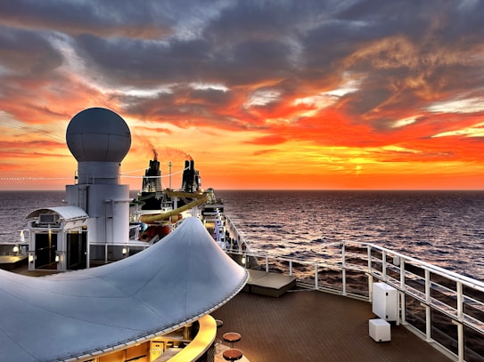 A cruise ship deck overlooking a vast ocean during sunset. The sky is ablaze with vibrant shades of orange, red, and yellow, blending into the darker hues of approaching night. A large radar sphere is prominently featured in the foreground, and the ship's layout includes seating and a water slide. Smoke trails rise from dual exhaust stacks, adding to the serene yet dynamic scene.