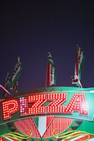 Bright marquee sign with the word 'Pizza' illuminated by red lights. The sign is adorned with geometric patterns and Italian flag colors. Several flags wave above the sign against a dark evening sky.