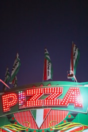 Bright marquee sign with the word 'Pizza' illuminated by red lights. The sign is adorned with geometric patterns and Italian flag colors. Several flags wave above the sign against a dark evening sky.