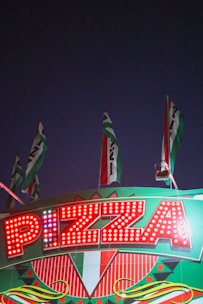 Bright marquee sign with the word 'Pizza' illuminated by red lights. The sign is adorned with geometric patterns and Italian flag colors. Several flags wave above the sign against a dark evening sky.