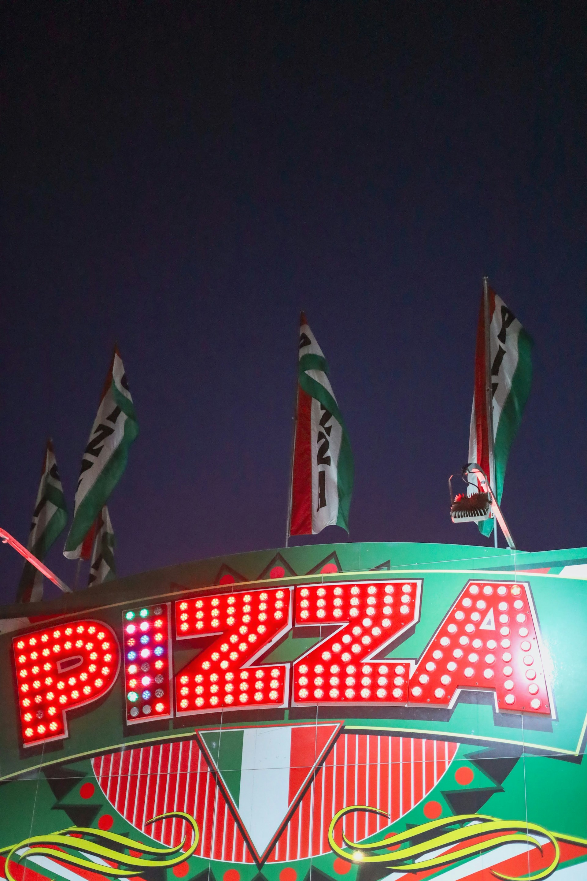 Bright marquee sign with the word 'Pizza' illuminated by red lights. The sign is adorned with geometric patterns and Italian flag colors. Several flags wave above the sign against a dark evening sky.