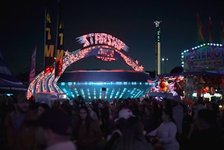 A bustling carnival scene at night, with a brightly lit spaceship-themed ride called 'Starship 4000' as the centerpiece. Colorful lights and signs illuminate the area, surrounded by a large crowd of people. Nearby, a 'Water Gun Fun' booth and a row of stuffed animal prizes add to the lively atmosphere.