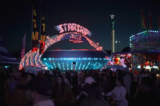 A bustling carnival scene at night, with a brightly lit spaceship-themed ride called 'Starship 4000' as the centerpiece. Colorful lights and signs illuminate the area, surrounded by a large crowd of people. Nearby, a 'Water Gun Fun' booth and a row of stuffed animal prizes add to the lively atmosphere.