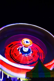 A brightly lit amusement park ride spinning at night, creating mesmerizing circular light trails in red, orange, blue, and yellow against a dark background. A tent with green and yellow stripes is partially visible at the bottom of the image.