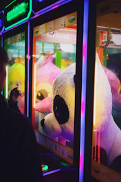 A large, plush panda toy is visible through the glass of a colorful arcade claw machine. Neon lights in various colors illuminate the machine, creating a vibrant, playful atmosphere. A person is standing in front of the machine, holding a coin or token.