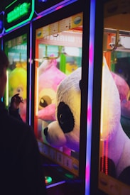 A large, plush panda toy is visible through the glass of a colorful arcade claw machine. Neon lights in various colors illuminate the machine, creating a vibrant, playful atmosphere. A person is standing in front of the machine, holding a coin or token.