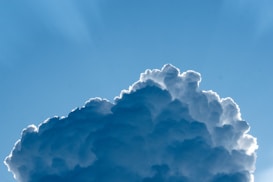 A large cumulus cloud with fluffy, white tops and a darker base against a clear blue sky. The sunlight creates a silver lining effect around the edges of the cloud.