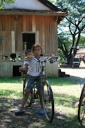 A young child balances on an old bicycle with another child seated on the back, both appearing in a rural setting. A wooden house with a thatched roof and a tree-filled background provide the backdrop, casting dappled sunlight on the ground.