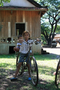 A young child balances on an old bicycle with another child seated on the back, both appearing in a rural setting. A wooden house with a thatched roof and a tree-filled background provide the backdrop, casting dappled sunlight on the ground.