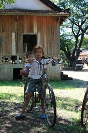 A young child balances on an old bicycle with another child seated on the back, both appearing in a rural setting. A wooden house with a thatched roof and a tree-filled background provide the backdrop, casting dappled sunlight on the ground.