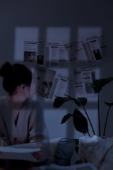 A person is sitting indoors, with their face turned towards a wall covered in newspaper clippings. The dim lighting casts a blue hue over the room, creating a contemplative atmosphere. A plant is visible next to the person, adding greenery to the setting.