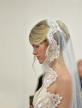 A bride is wearing a white lace veil and an elegant wedding dress. Her hair is styled in an updo with a loose strand flowing down, adding a romantic touch. The intricate lace detail of the veil frames her face delicately.