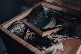 A wicker box filled with various nostalgic items, including a porcelain doll's head, a closed leather-bound book, a wooden picture frame, and dried flowers. Soft light highlights the textures and creates shadows, adding a mysterious atmosphere.