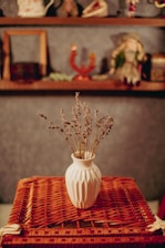 A white ceramic vase filled with dried lavender is placed on a textured, woven surface. In the background, there are shelves with various decorative objects including a doll and small ornaments.