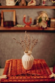 A white ceramic vase filled with dried lavender is placed on a textured, woven surface. In the background, there are shelves with various decorative objects including a doll and small ornaments.