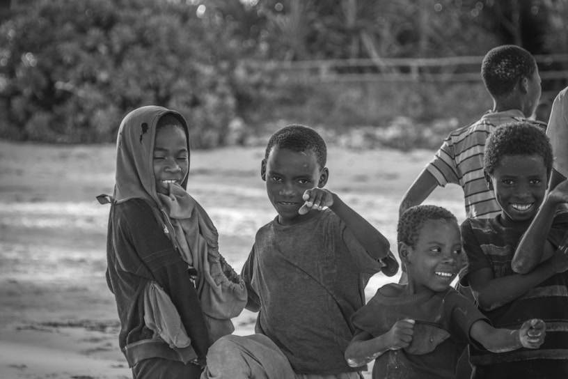 A group of children smiling and playing together on a sandy area, with one child wearing a headscarf. They appear to be enjoying each other's company.