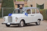 A classic white car, resembling a vintage taxi, is adorned with blue ribbon decorations on its front and side mirrors. It is parked on a gravel surface with greenery and an old building in the background.