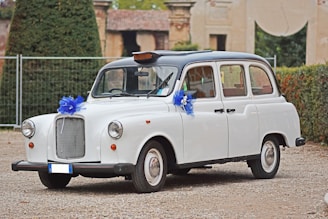 A classic white car, resembling a vintage taxi, is adorned with blue ribbon decorations on its front and side mirrors. It is parked on a gravel surface with greenery and an old building in the background.